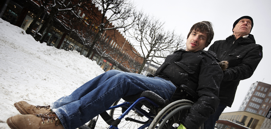 En medelålders, manlig personlig assistent kör en yngre manlig brukare i rullstol på ett snötäckt torg.