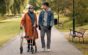 &Auml;ldre kvinna med rollator g&aring;r med ung man i park.
