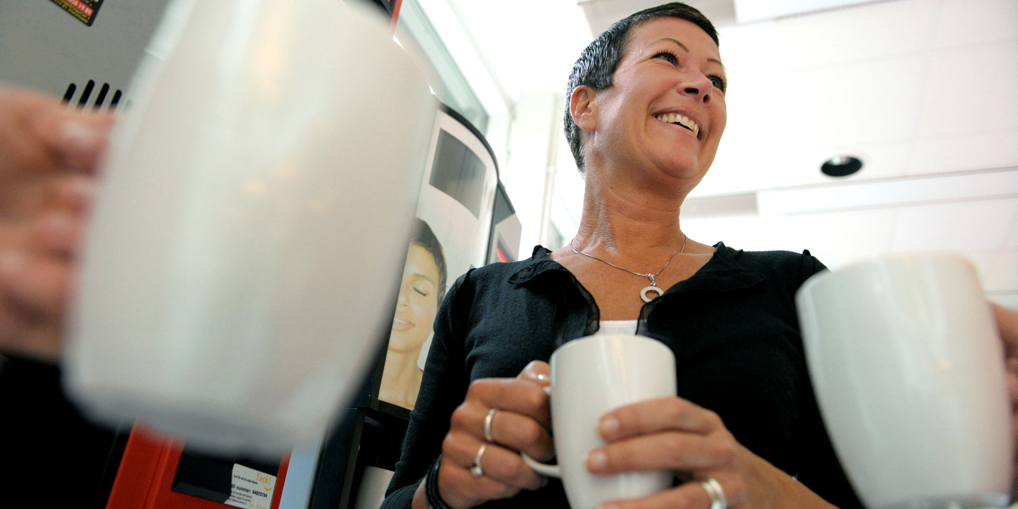 Leende kvinna med kaffemugg. Runt henne flera andra kaffemuggar som h&aring;lls av h&auml;nder.