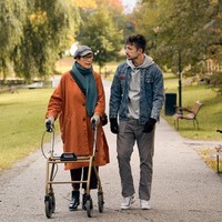 Äldre kvinna med rullator och yngre man promenerar i en park om hösten. 