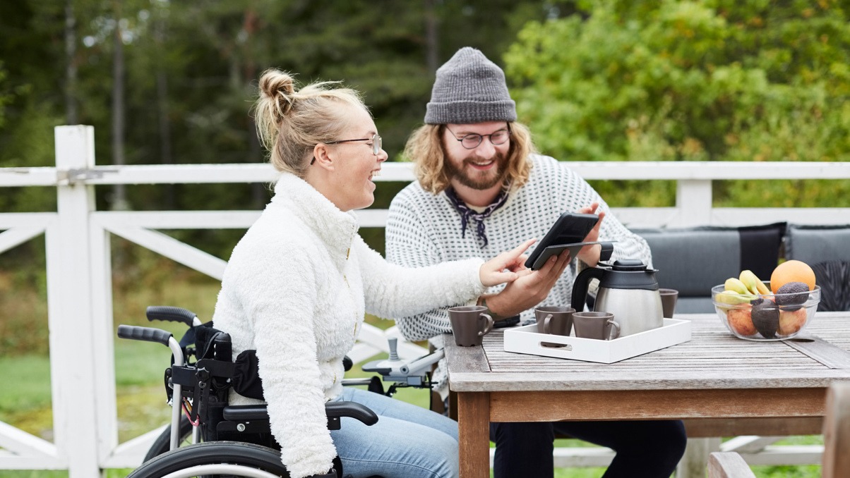 En man som jobbar som personlig assistent tittar p&aring; en l&auml;splatta tillsammans med en ung kvinna i rullstol.