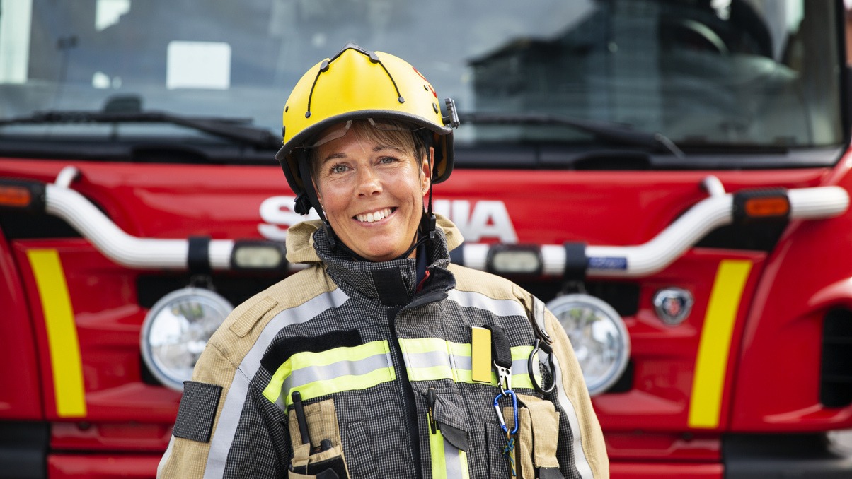 Kvinnlig brandman som ler och st&aring;r framf&ouml;r en brandbil.