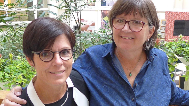 susanne hylander, upphandlare, och agneta kallstenius, arbetsmilj&ouml;controller, ler mot kameran och st&aring;r t&auml;tt ihop i en ljushall med gr&ouml;nska bakom ryggen.