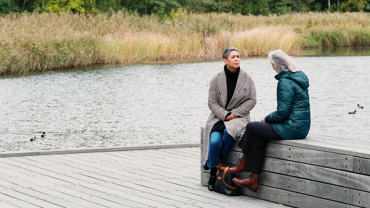 Tv&aring; kvinnor sitter p&aring; en brygga intill ett vattendrag.
