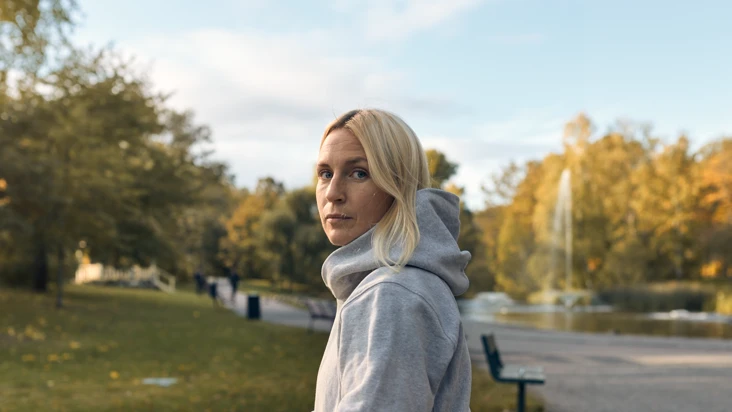 En kvinna i gr&aring; huvtr&ouml;ja g&aring;r utomhus och tittar in i kameran.