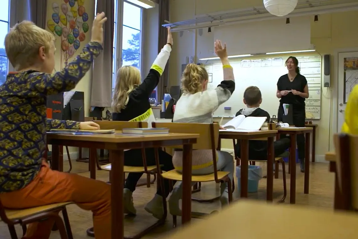 En grupp barn som sitter vid sina b&auml;nkar i ett klassrum.